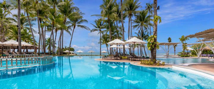 Leuchtend blauer Pool im Fokus, umgeben von Palmen und Liegen mit Sonnenschirmen, bei schönem Wetter mit vereinzelten Wolken.